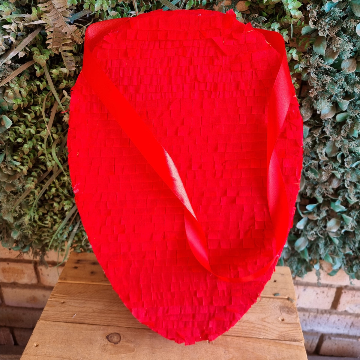 A bright red woven pinata shaped like a stylized face, displayed on a wooden table in front of a green leafy hedge background.