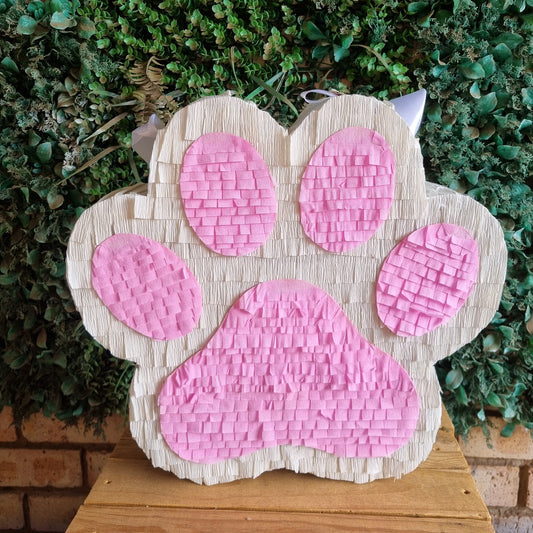A cat OR DOG  paw print piñata decoration with pink woven details on a beige wood-grain base, displayed on a wooden surface against a green foliage background.