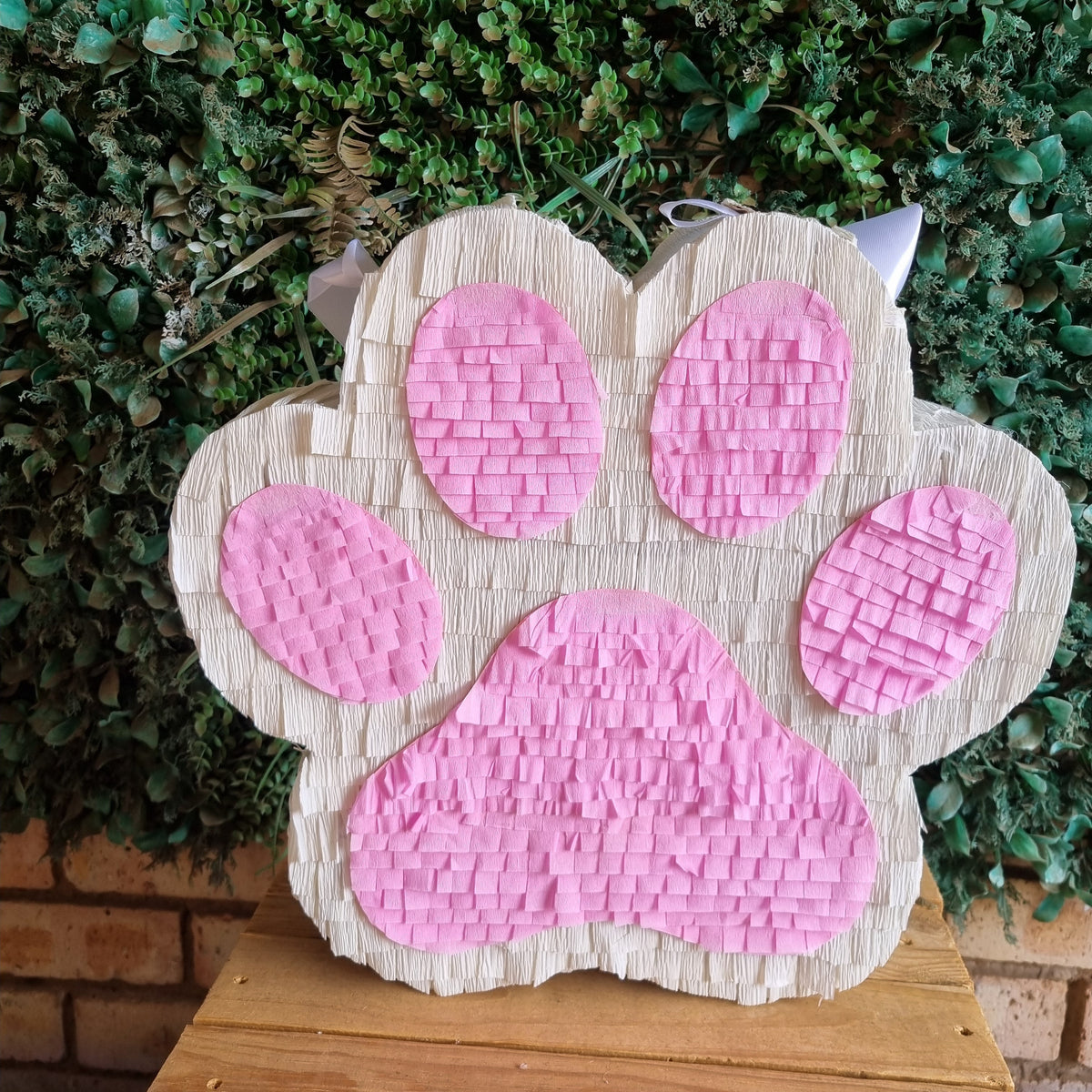 A paw-shaped pinata with pink paw pads and small white cat ears, displayed on a wooden table in front of a green leafy wall.