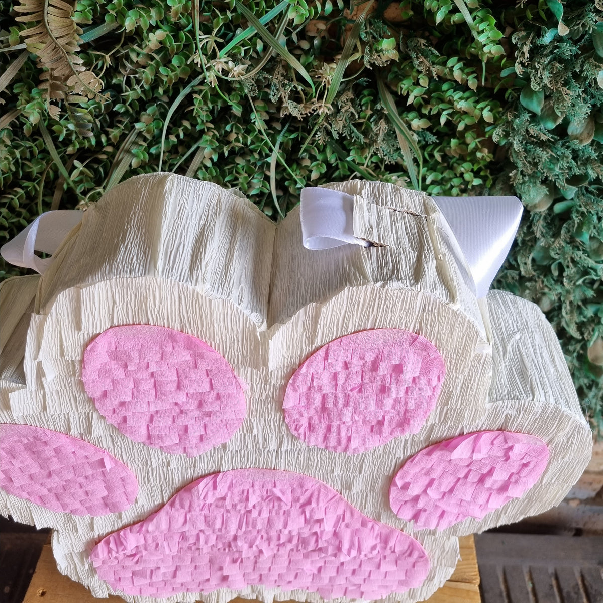 A novelty piñata shaped like a cat paw, made of textured white tissue paper with pink paw pads, photographed against a green foliage backdrop.