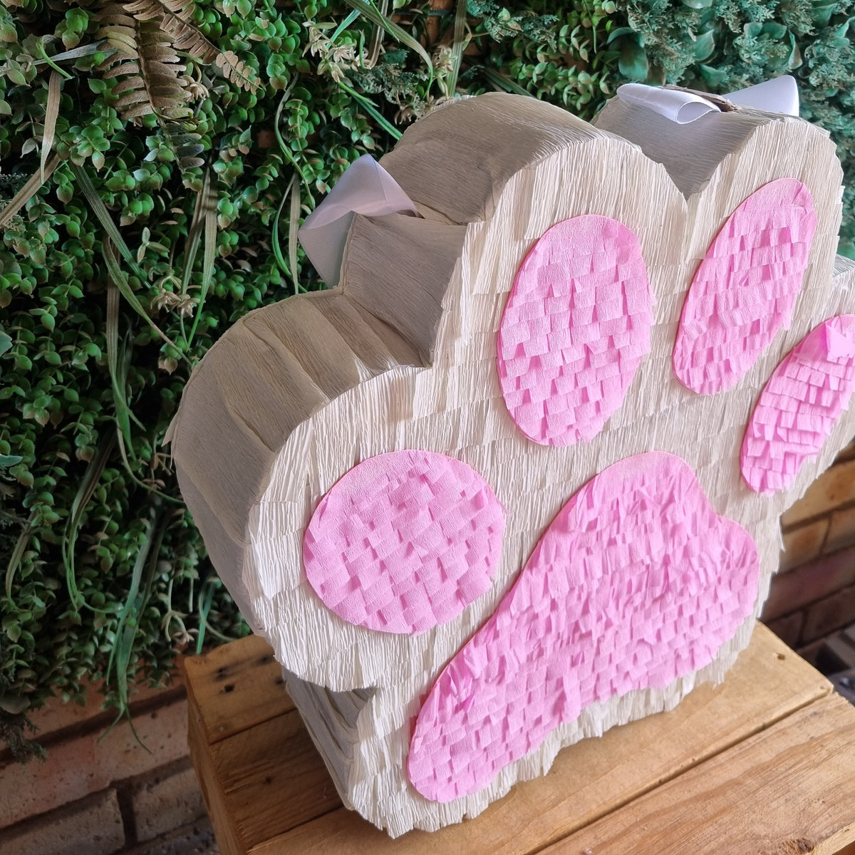A DOG OR cat paw print piñata party decoration with pink textured pads, displayed on a wooden surface in front of a green foliage background.