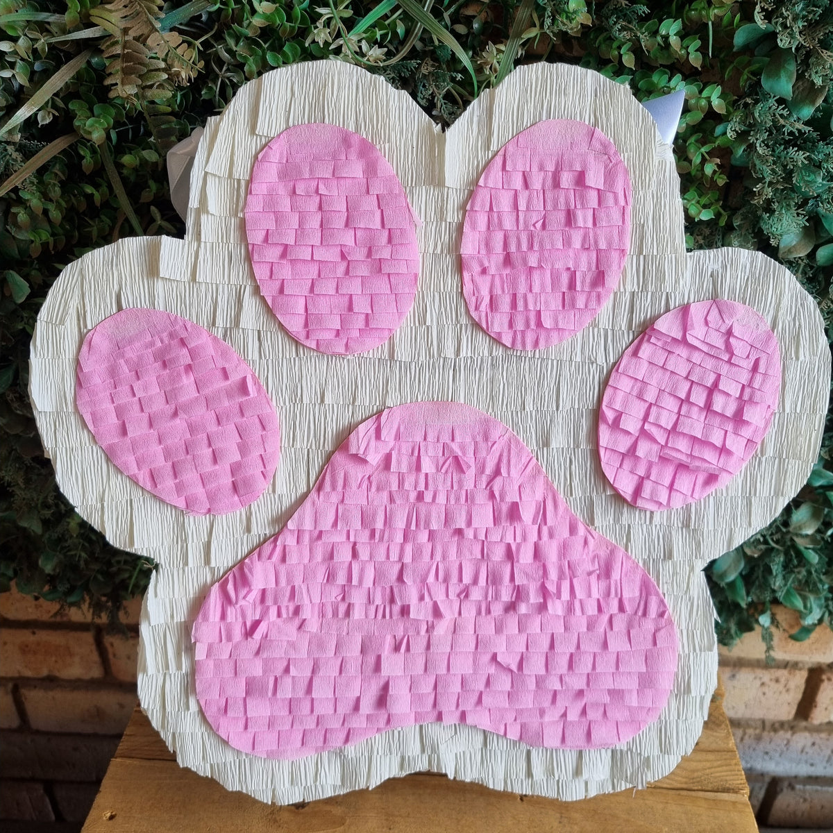 A  DOG OR cat paw print piñata party decoration made of white and pink crinkle-cut paper, displayed on a wooden table with green foliage in the background.