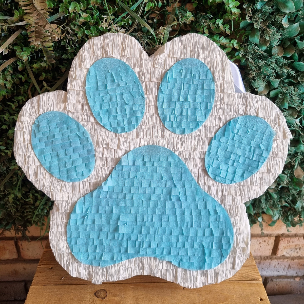 A paw print shaped party piñata with light blue textured centers and a beige fringed paper border, displayed on a wooden surface against a green leafy background.