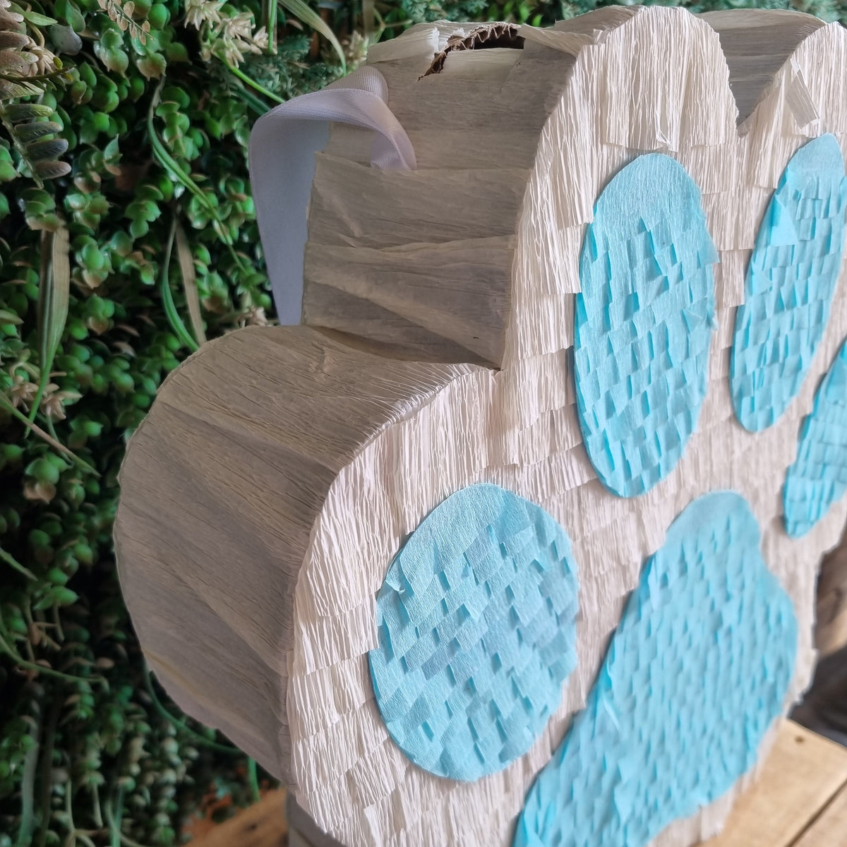 A  piñata shaped like a paw print, featuring textured light wood sides and bright blue circular patterned inserts on the paw pads, displayed on a wooden surface with green foliage in the background.