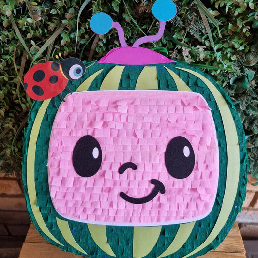 A cute watermelon-themed pinata with a smiling pink face, green and yellow stripes, a purple antenna, and a ladybug accent, displayed on a wooden surface against a green foliage background.