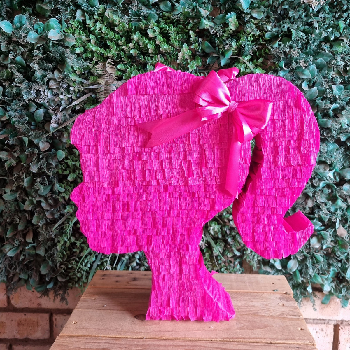 A bright pink Barbie-style hair silhouette piñata decoration with a matching bow, displayed on a wooden surface against a green leafy wall background.