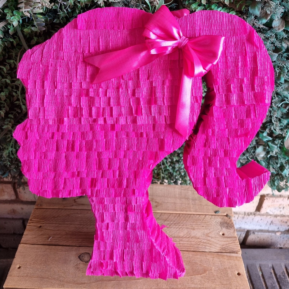 A bright pink elephant-shaped pinata with a textured paper weave pattern and a matching pink satin bow on its back, displayed on a wooden surface against a green leafy background.