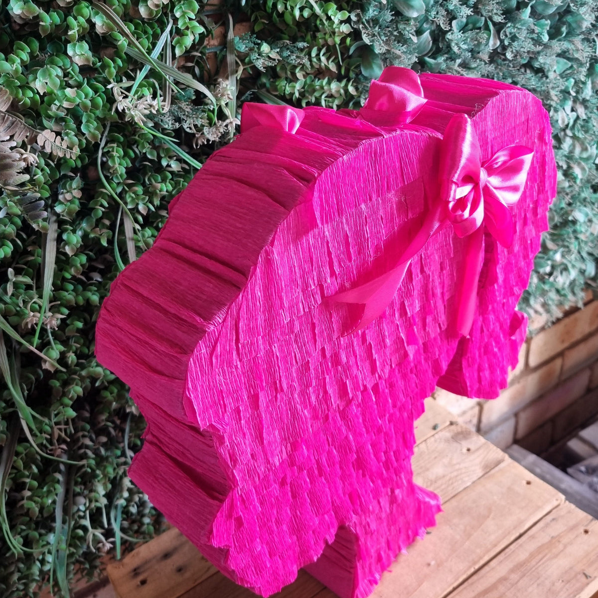 A bright pink elephant-shaped paper piñata party decoration with a matching bow, displayed on a wooden crate in front of a green leafy wall.