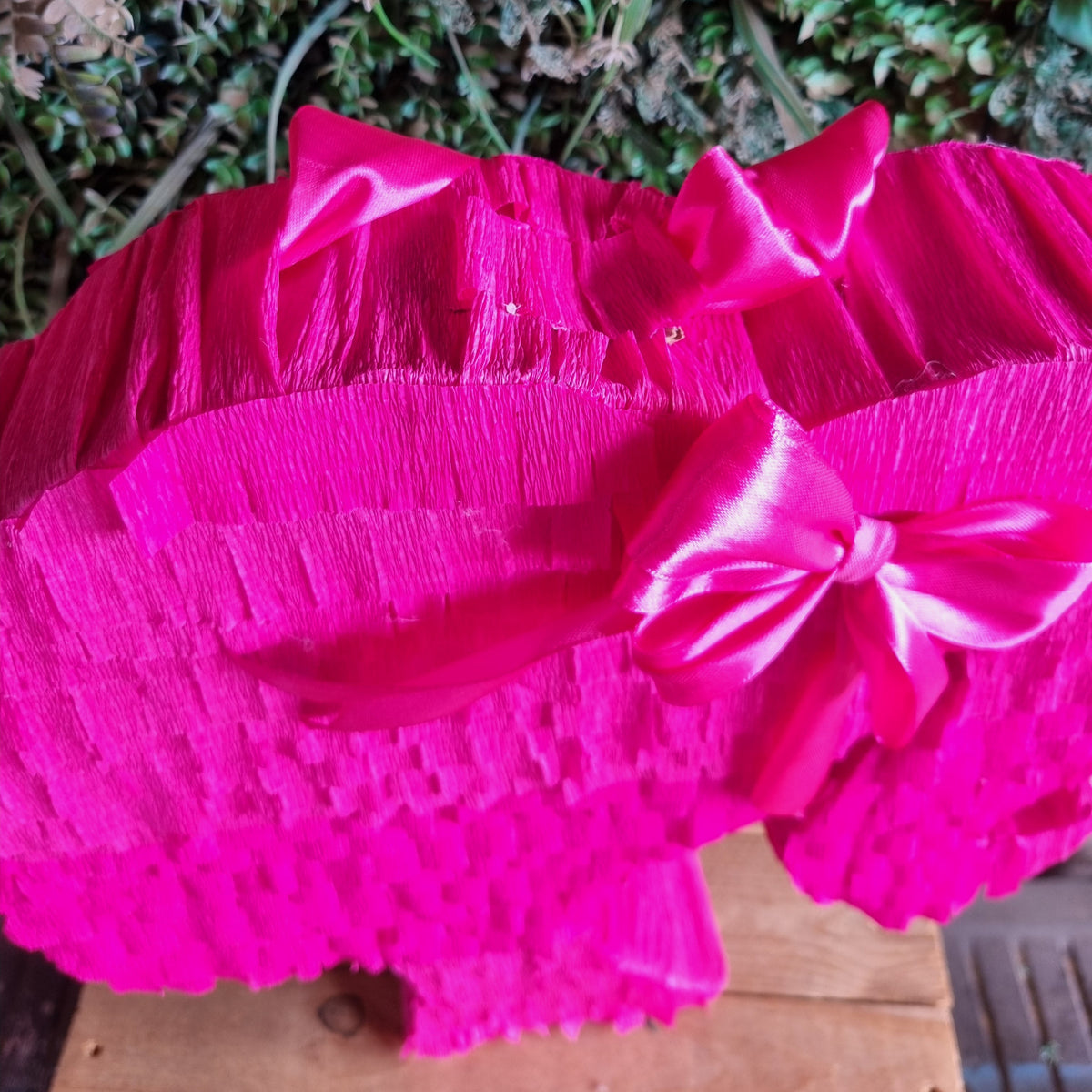 A close-up of a bright hot pink ruffled fabric accessory with large satin bows, photographed against a background of green foliage and a wooden surface.