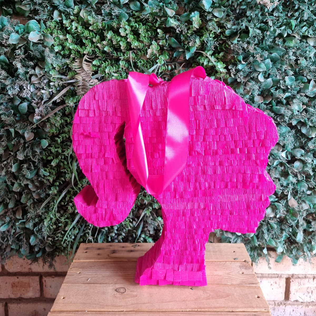 A bright pink Barbie-themed pinata shaped like a woman's profile with a glossy bow, displayed on a wooden table in front of a green leafy wall.