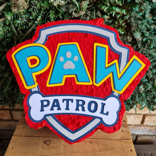 A red, blue, and yellow Paw Patrol themed pinata sign displayed on a wooden surface in front of a green leafy background.