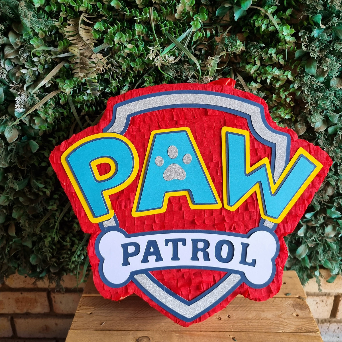 A red Paw Patrol themed pinata shaped like the official Paw Patrol shield, featuring blue 'PAW' letters with yellow outlines and a white bone reading 'PATROL' underneath, displayed on a wooden surface in front of a green foliage backdrop.