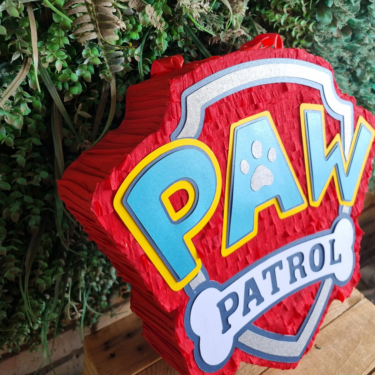 A red Paw Patrol themed party piñata sign featuring the PAW Patrol logo, displayed in front of a green foliage backdrop.
