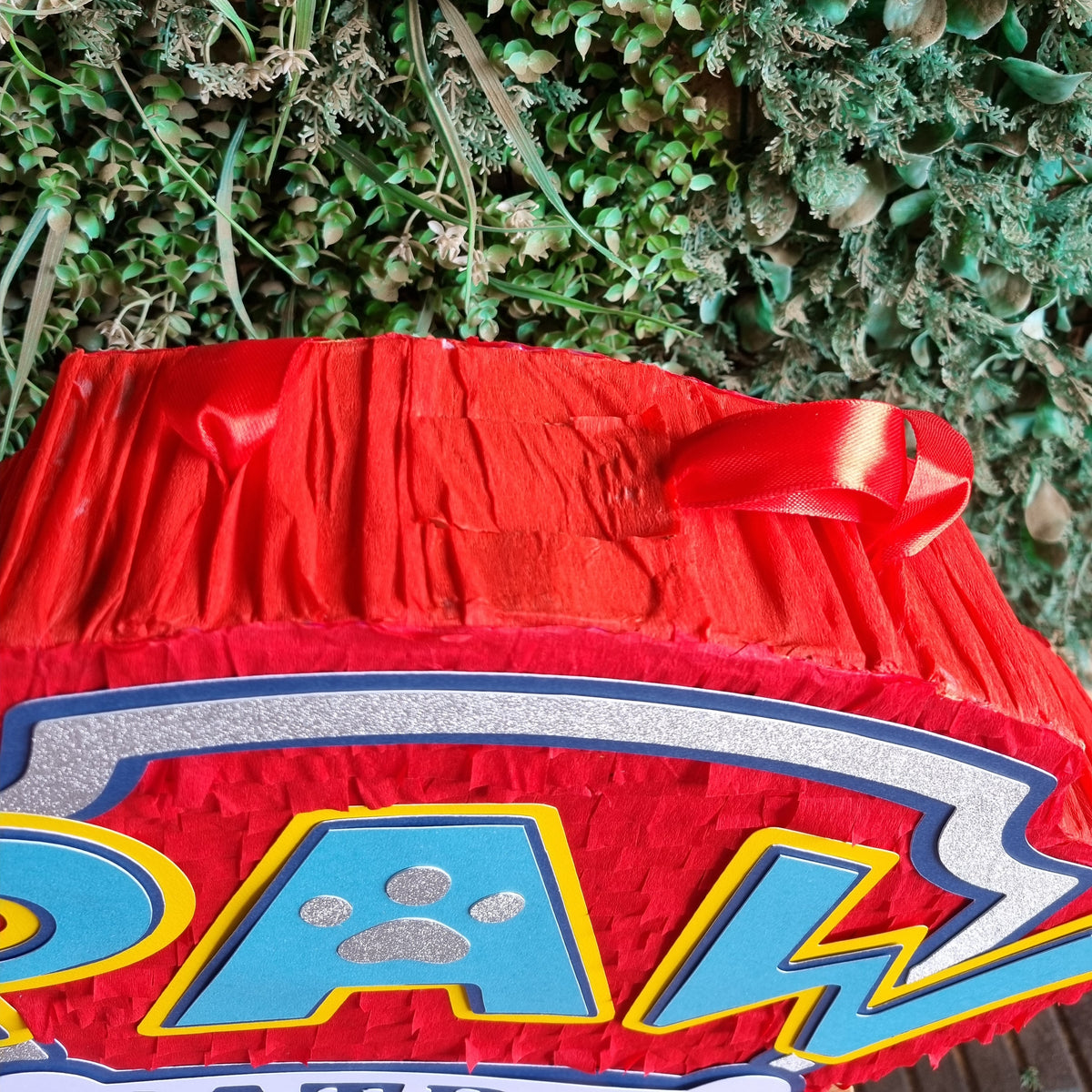 A close-up of a red and blue 'PAW' themed party decoration or banner, featuring a red pleated top with a ribbon bow and a green foliage backdrop.
