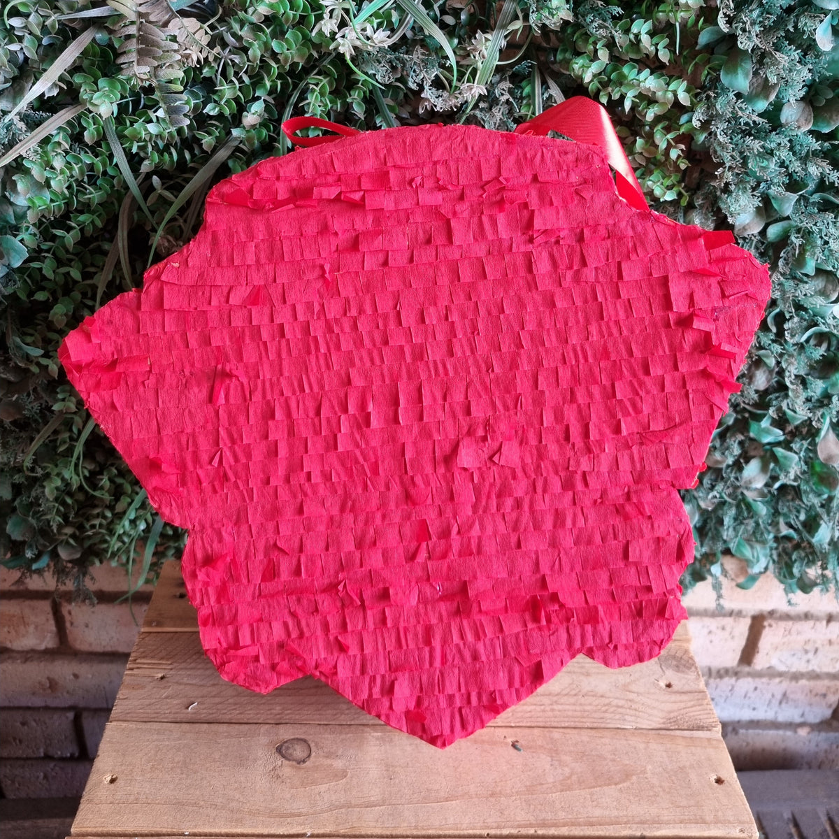 A bright red paper piñata shaped like a strawberry, displayed on a wooden table in front of a green leafy wall backdrop.