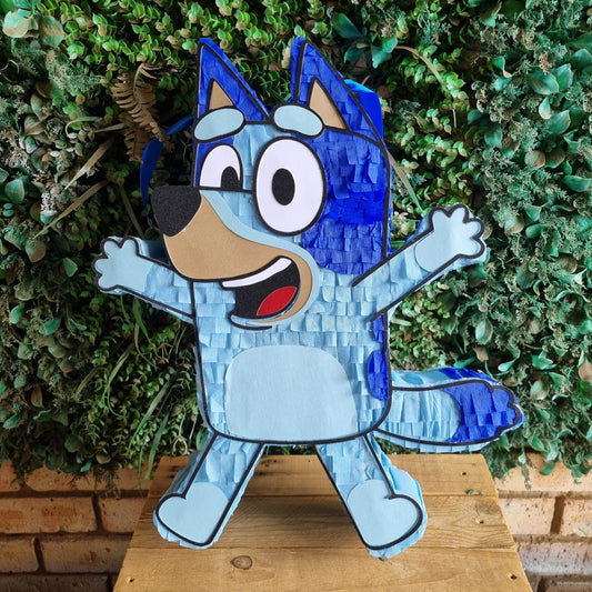 A blue cartoon dog piñata cutout, styled like Bluey, standing on a wooden table in front of a green leafy wall.