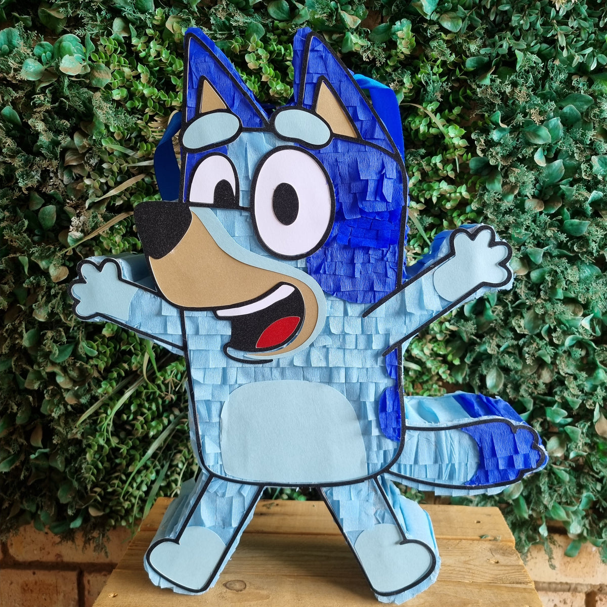 A Bluey character piñata shaped like Bingo, featuring a blue and light blue textured body with a tan snout and black outline, standing on a wooden surface against a green leafy background.