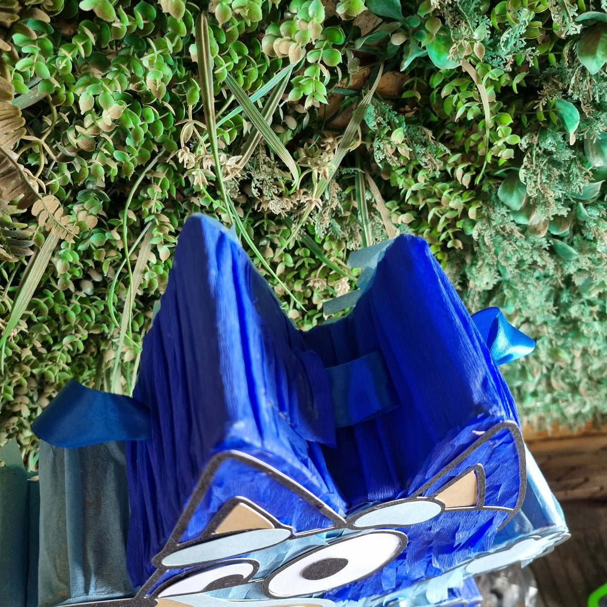 A blue cartoon wolf-shaped party decoration or piñata with a printed face, photographed against a green leafy backdrop and wooden surface.