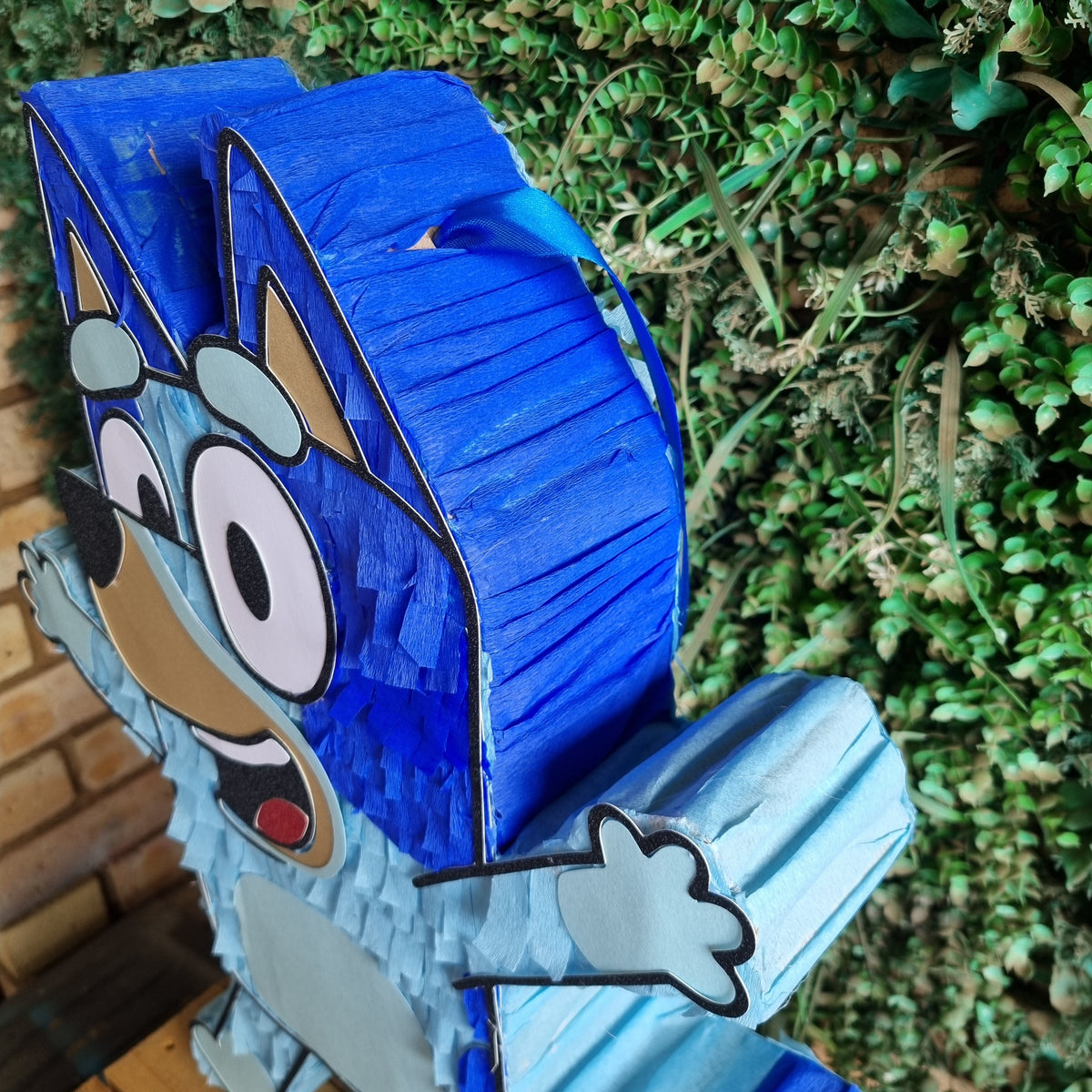 A blue piñata shaped like the Bluey character Bingo, featuring a textured paper finish and cartoon face details, set against a green foliage background.