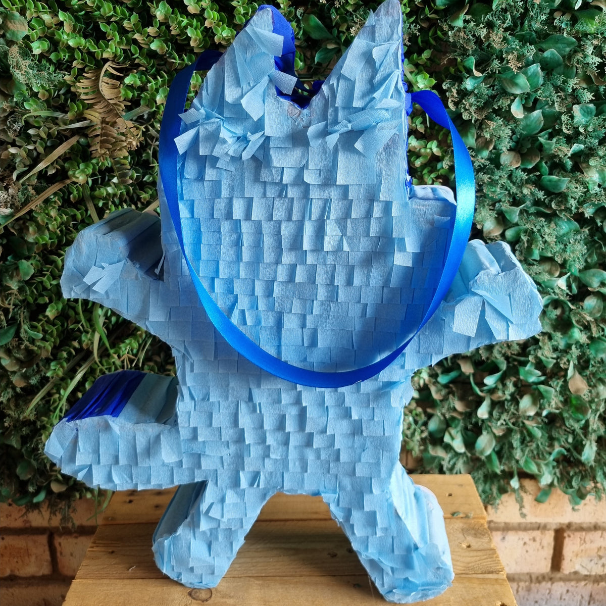 A blue paper bag shaped like a wolf or cat, standing on a wooden surface in front of a green leafy wall.