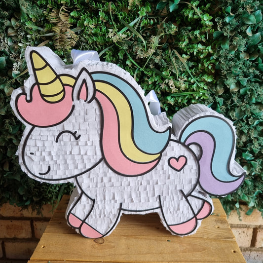A unicorn-shaped pinata with a pastel rainbow mane and tail, standing on a wooden surface against a green leafy background.