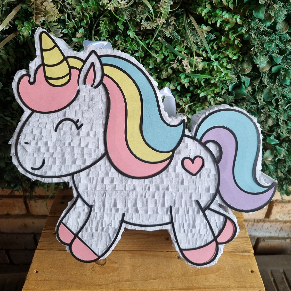 A white unicorn piñata party decoration with a pastel rainbow mane and tail, standing on a wooden surface against a green leafy background.