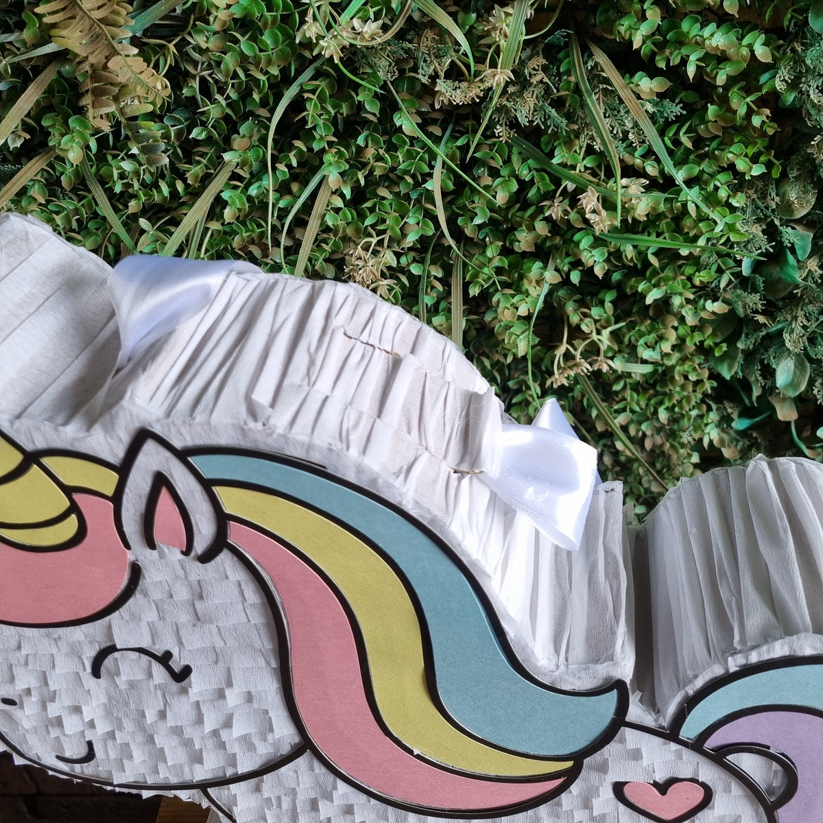 A close-up of a white pleated party hat featuring a colorful cartoon unicorn face with pink, yellow, and blue details, set against a background of green foliage.