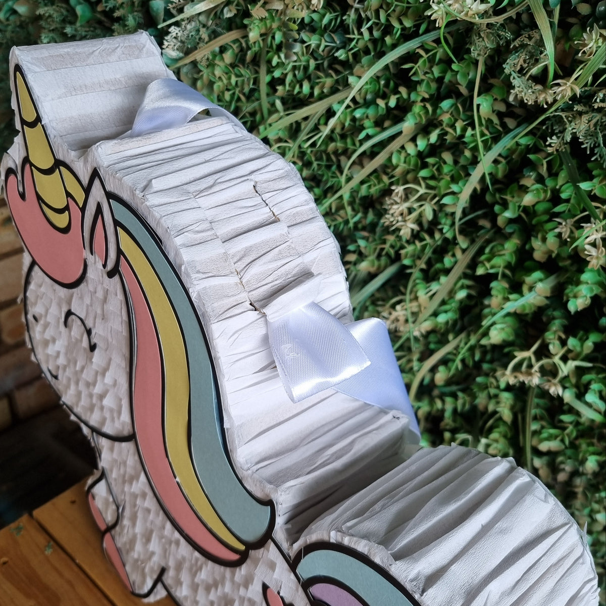 A large, white, crinkle-textured unicorn party hat or prop displayed on a wooden surface in front of a green foliage wall.