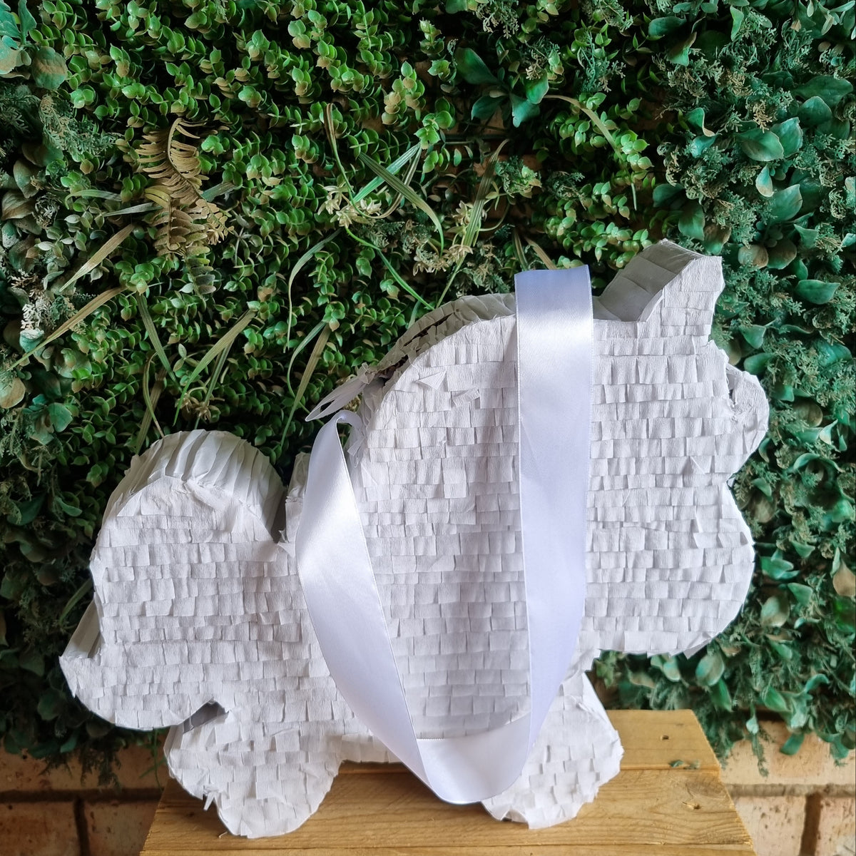 A white paper piñata shaped like a rabbit, decorated with a white satin ribbon handle, displayed on a wooden surface against a green leafy wall background.