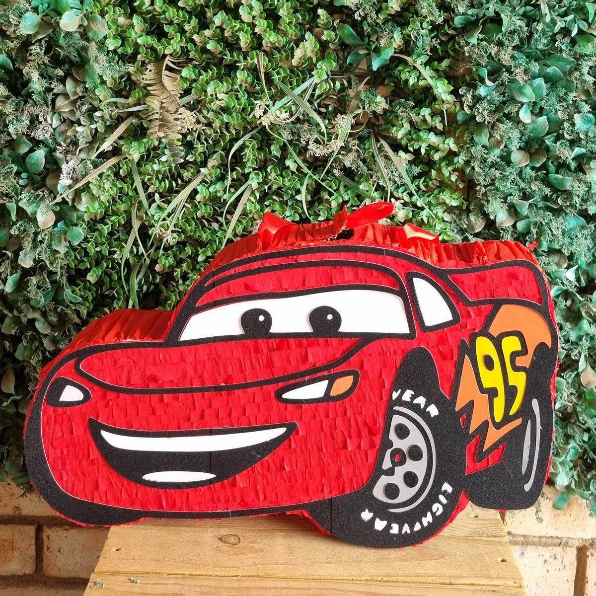 A red Lightning McQueen piñata shaped like the cartoon race car, displayed on a wooden stool in front of a green leafy wall.