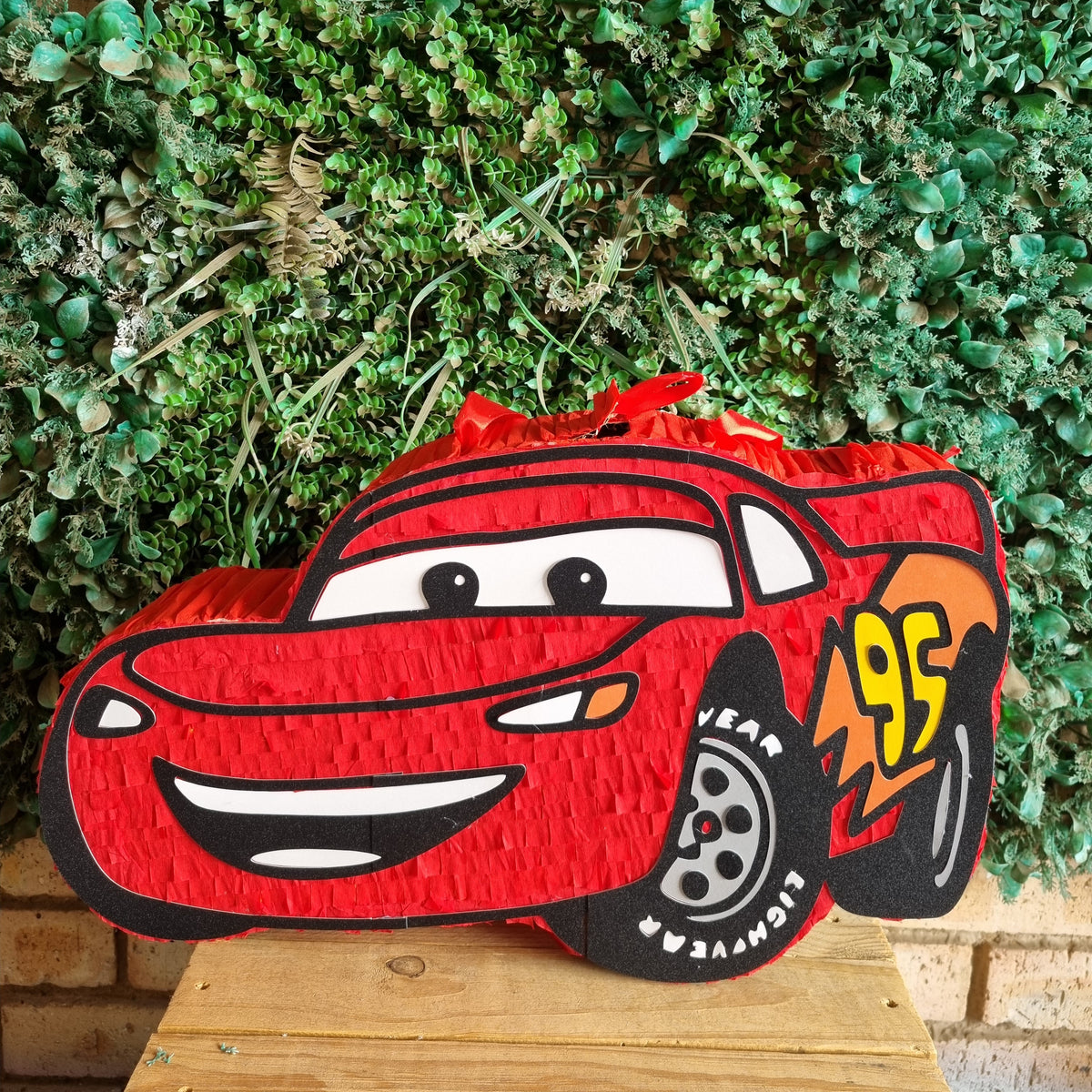 A red Lightning McQueen piñata shaped like the character car, displayed on a wooden surface in front of a green leafy wall.