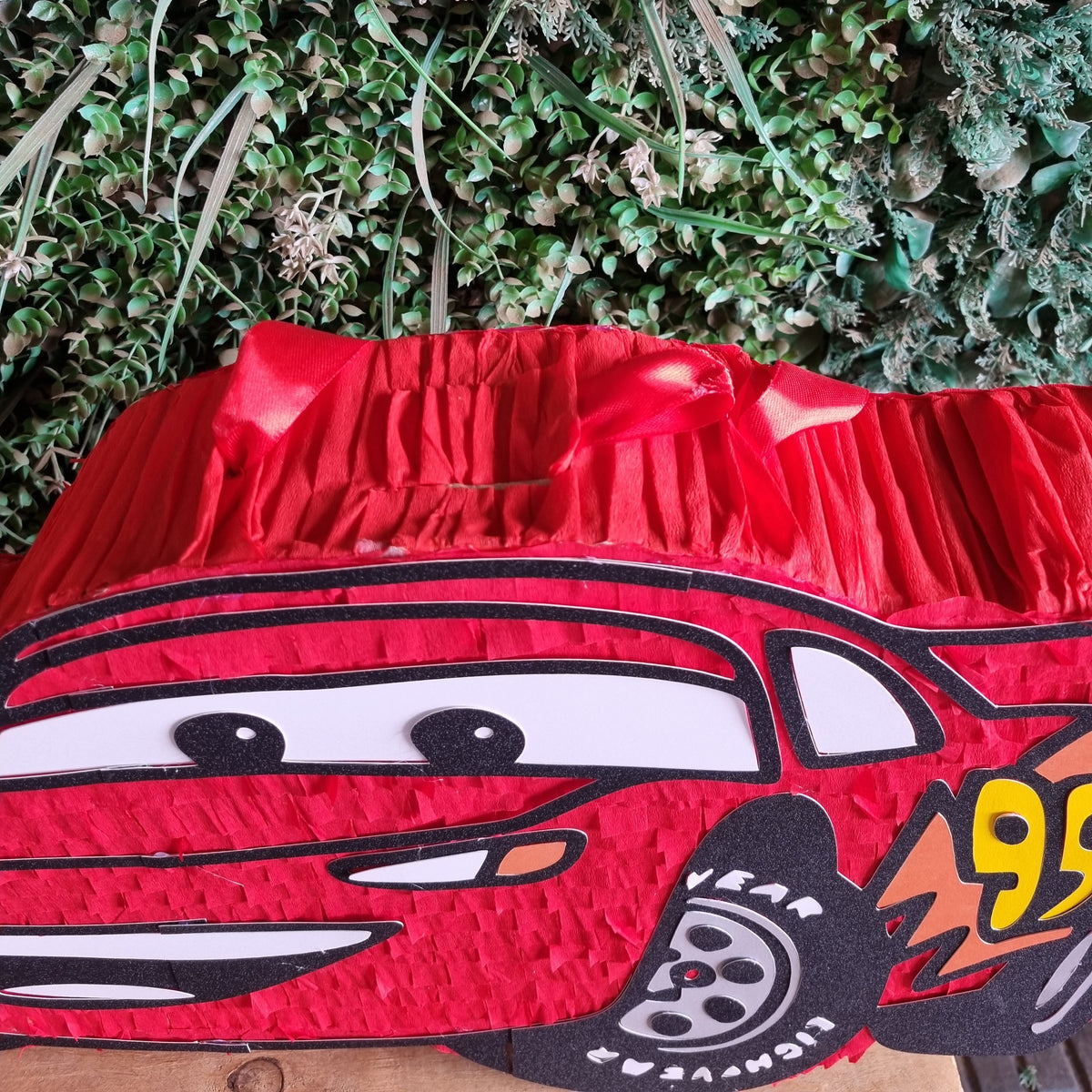 A red Lightning McQueen piñata party decoration with a black, white, and yellow printed design, set against a green foliage backdrop.