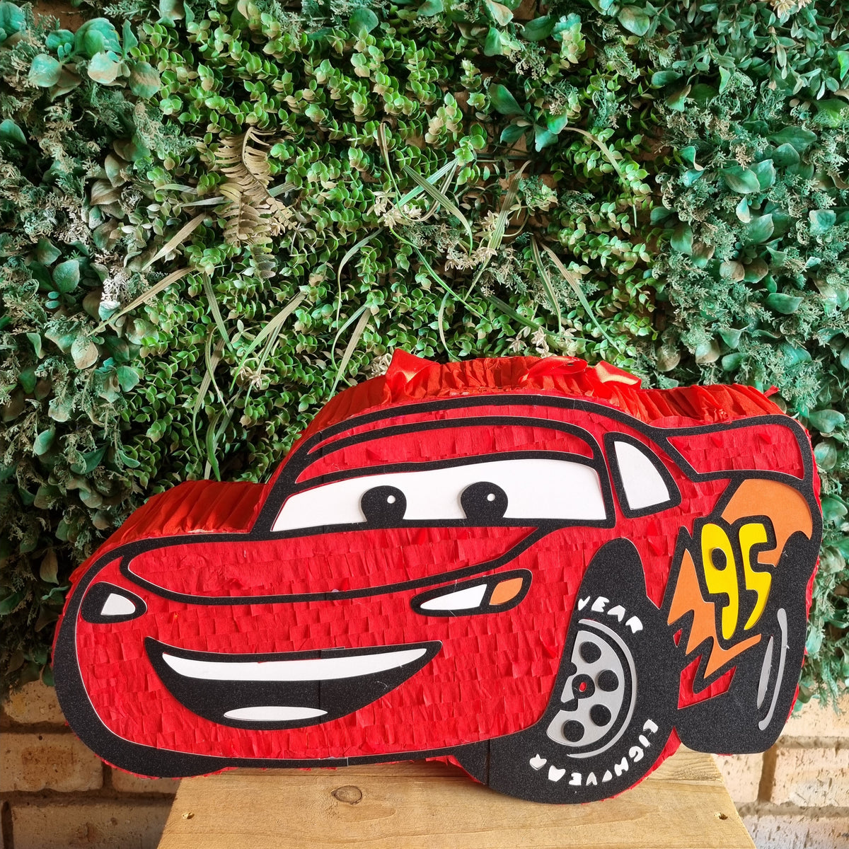 A red Lightning McQueen piñata shaped car decoration featuring his cartoon face and number 95, displayed on a wooden surface in front of a green leafy wall.