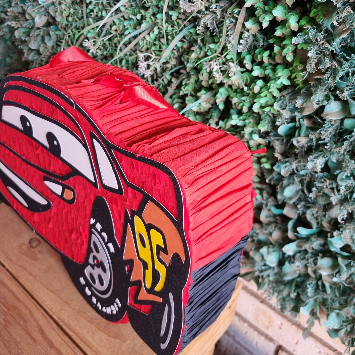 A red Lightning McQueen piñata party decoration shaped like his race car, displayed on a wooden surface in front of a green foliage backdrop.