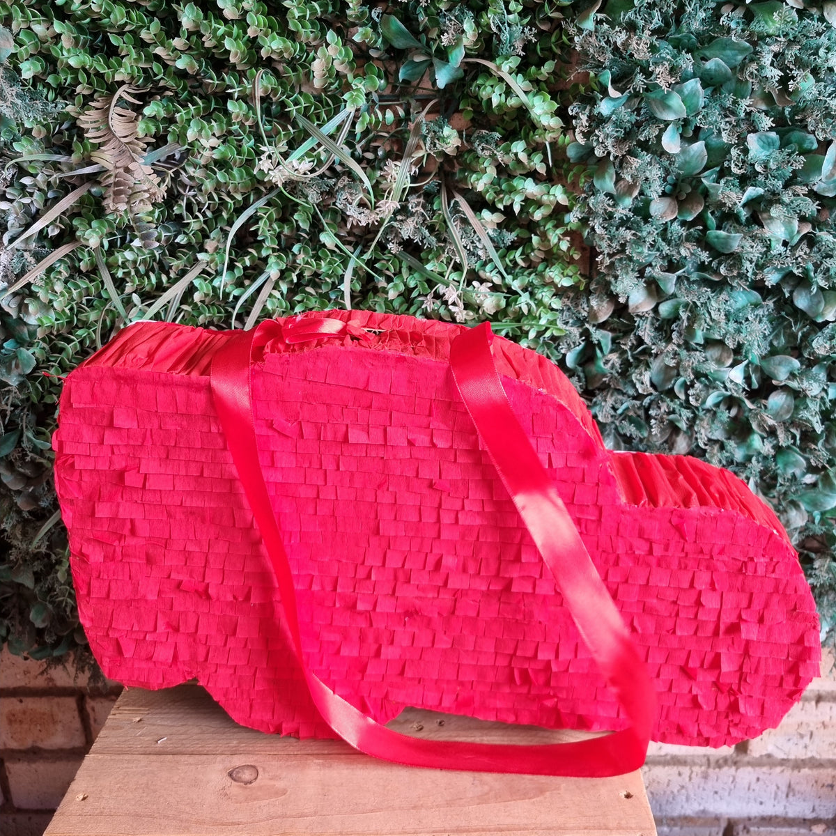 A bright red, cloud-shaped woven paper piñata bag with matching red shoulder straps, displayed on a wooden surface against a green ivy wall background.