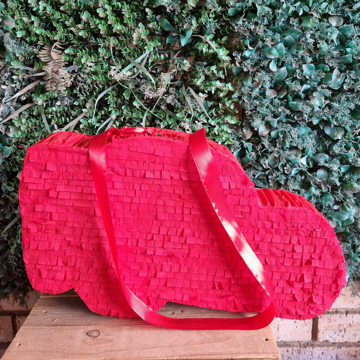 A bright red heart-shaped paper piñata with a matching red ribbon handle, displayed on a wooden surface in front of a green leafy wall.