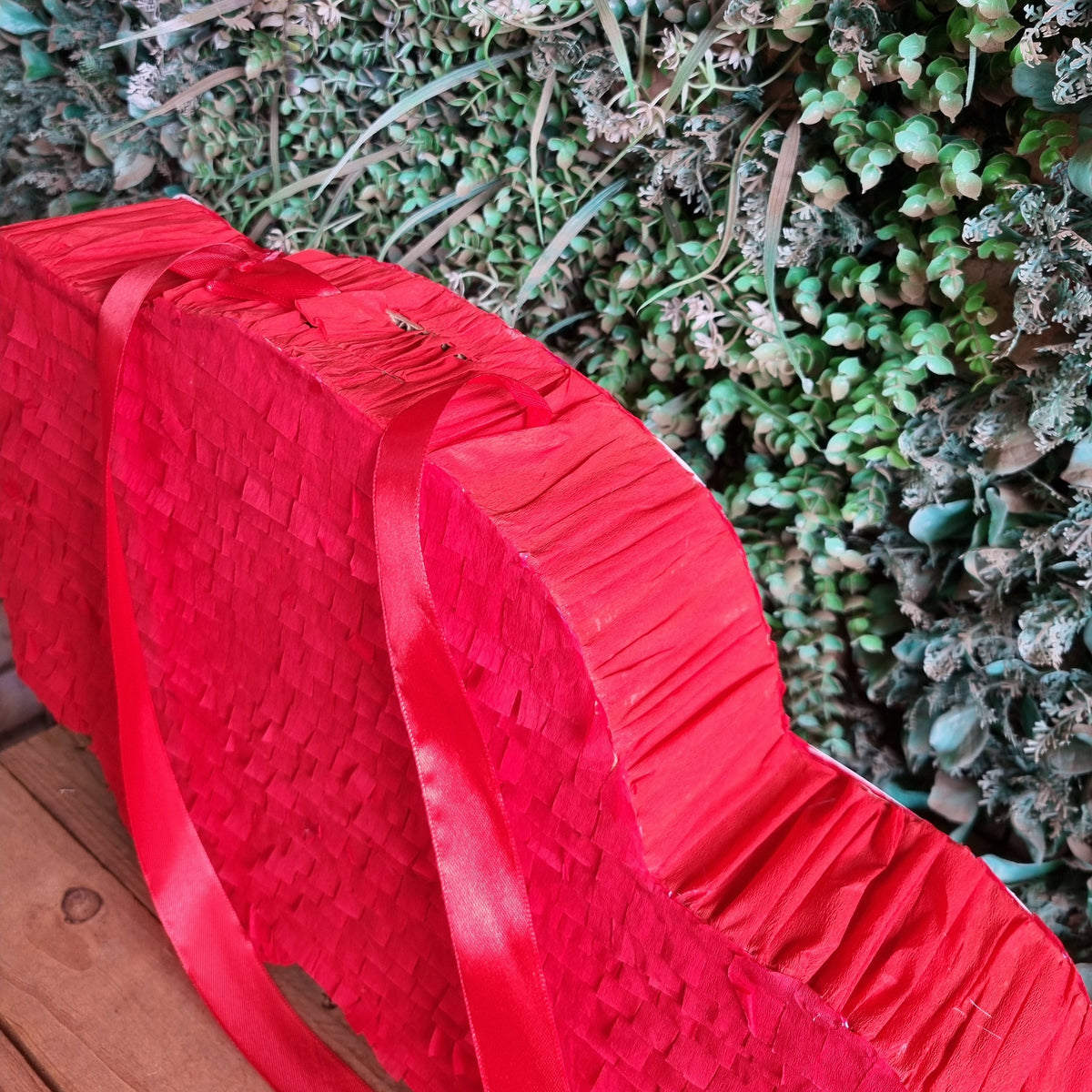 A red heart-shaped paper piñata with pleated texture and a long red ribbon handle, photographed outdoors against a green succulent wall and wooden surface.