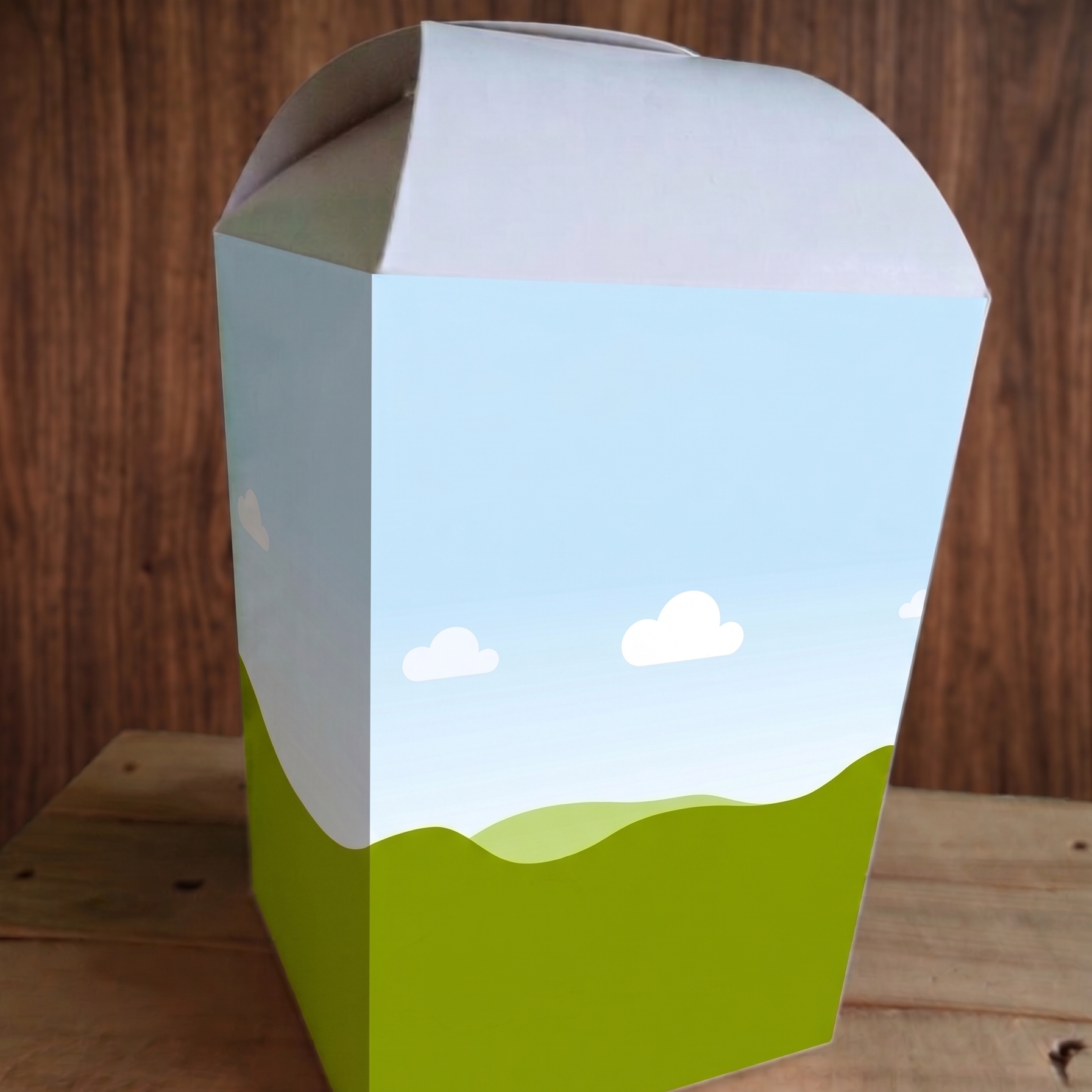 Cardboard box with a scenic design of a blue sky with clouds and green landscape on a wooden surface.