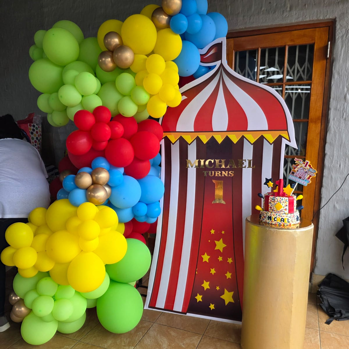 Colorful balloon arch with a circus-themed backdrop and cake on a stand.