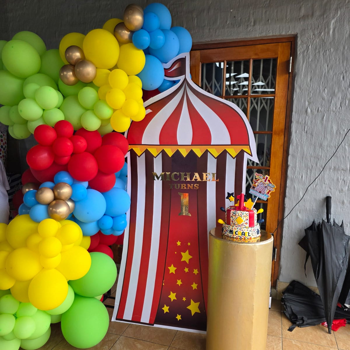 Colorful balloon display with a circus-themed backdrop and cake on a gold stand.