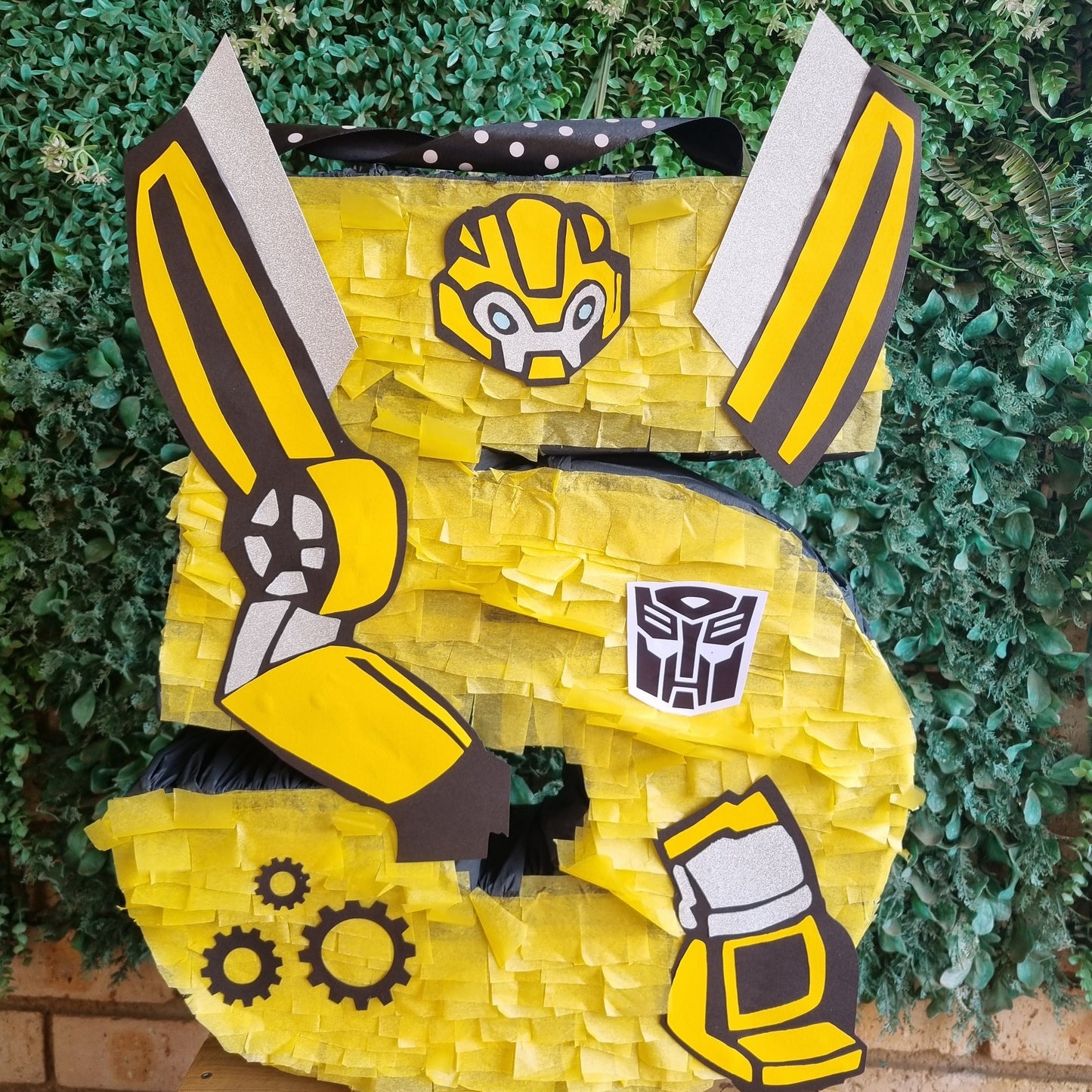 Yellow pinata shaped like a robot with black and gray accents against a green leafy background.