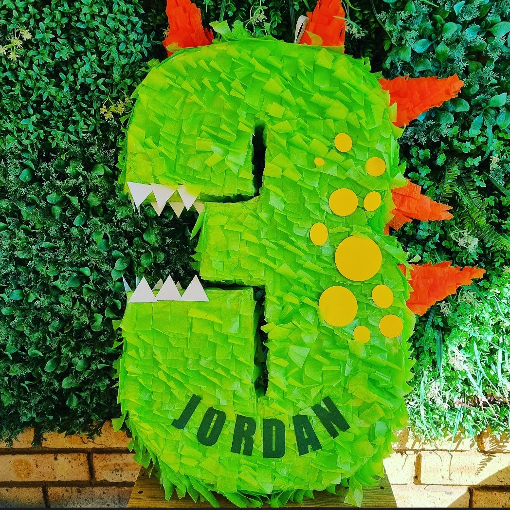 Green dinosaur-shaped piñata with 'JORDAN' on a green leafy background