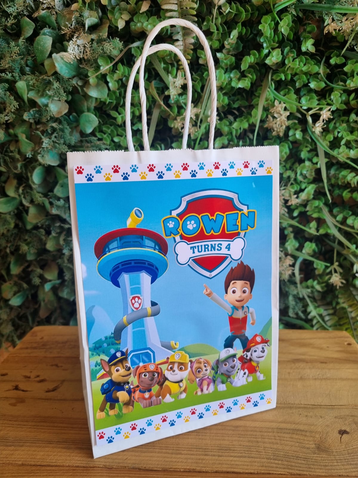 Gift bag with Paw Patrol design featuring a child and tower, against a green leafy background.